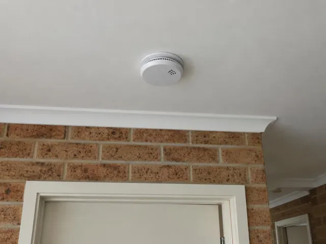 Ceiling smoke alarm above a brick hallway wall and door frame in Bateau Bay